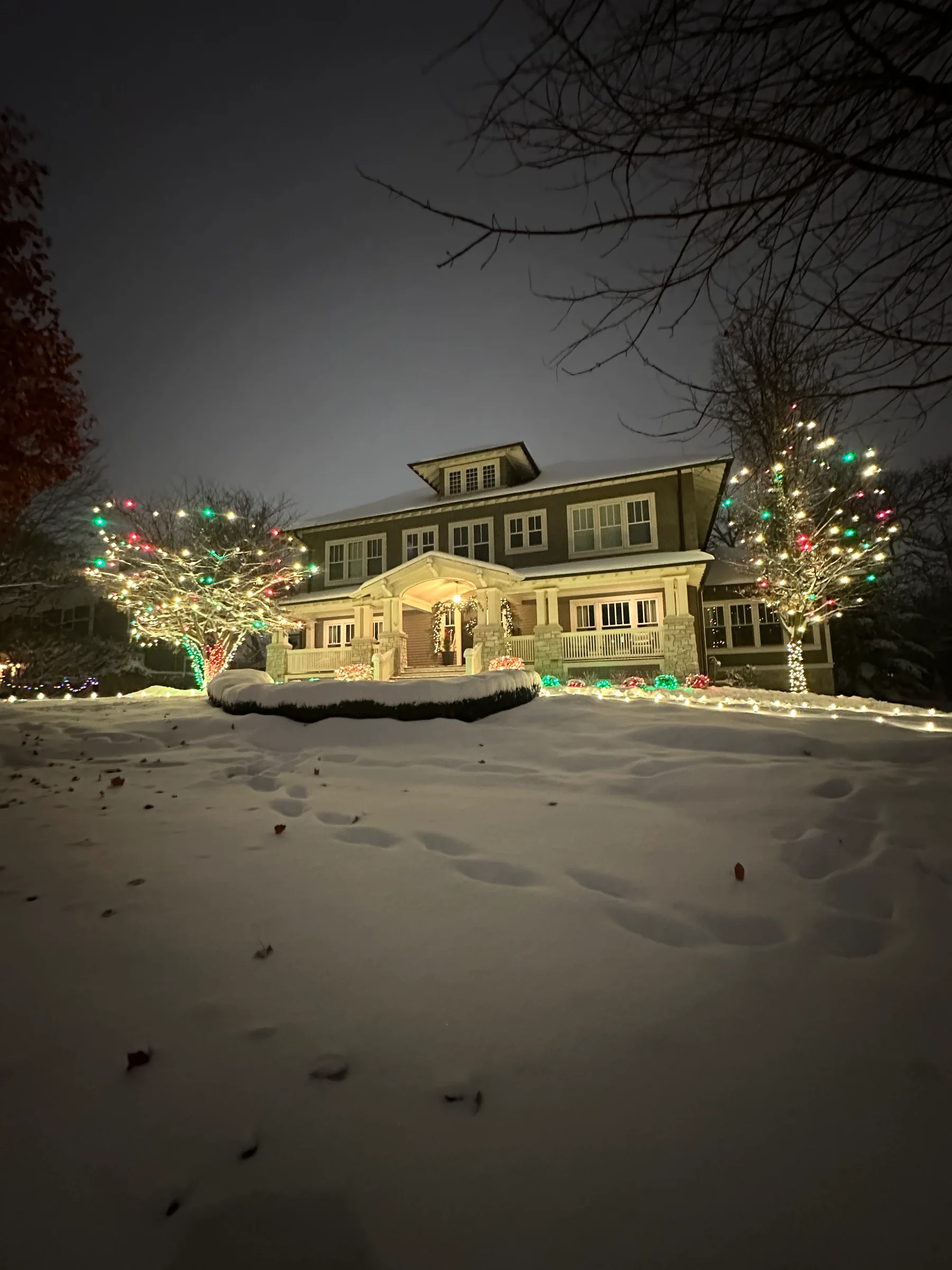 Snow-covered home with red, green, and warm-white lights across trees, landscaping, and the front entry