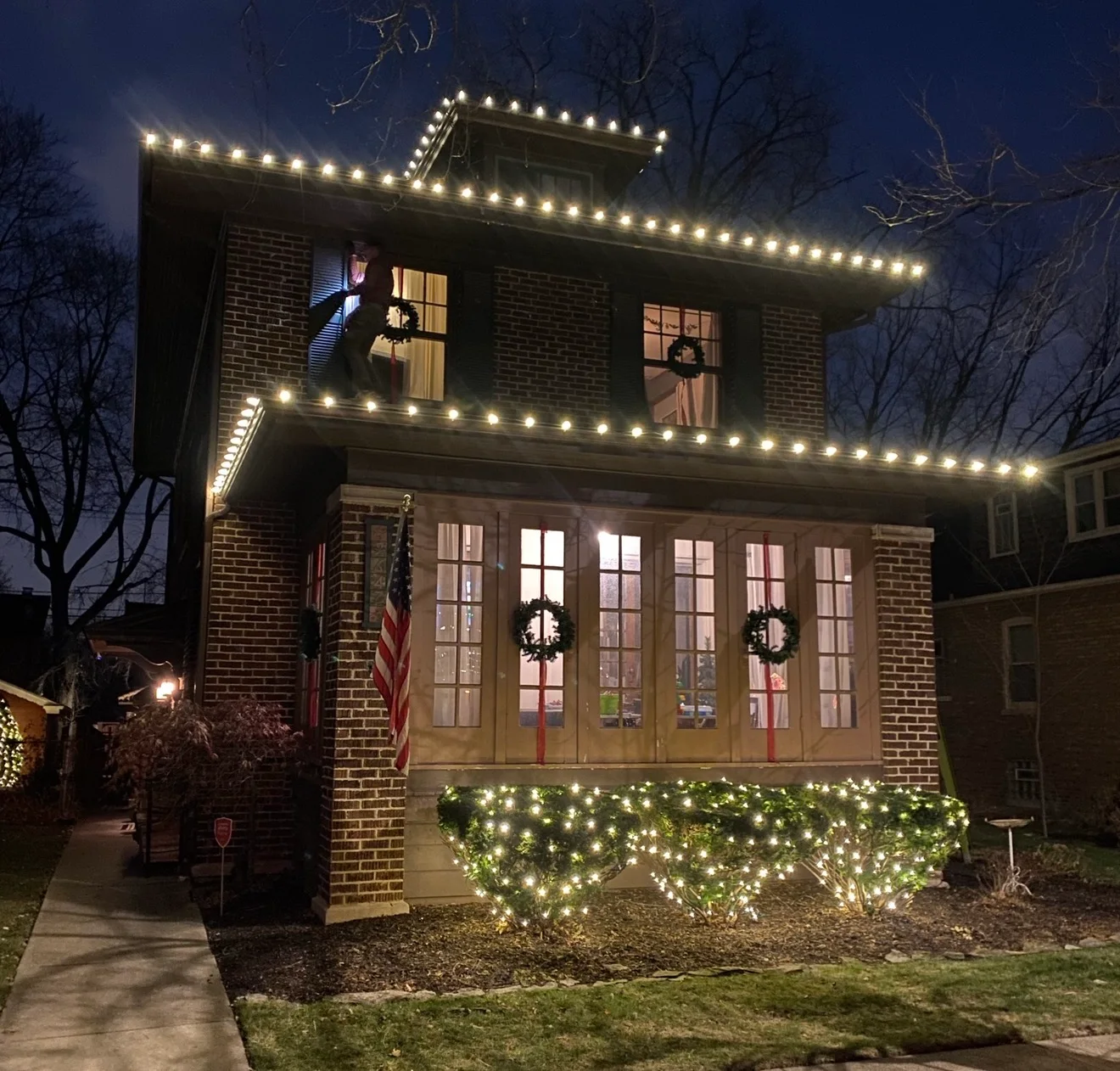 Two-story brick home with warm-white roofline, porch, and shrub lighting