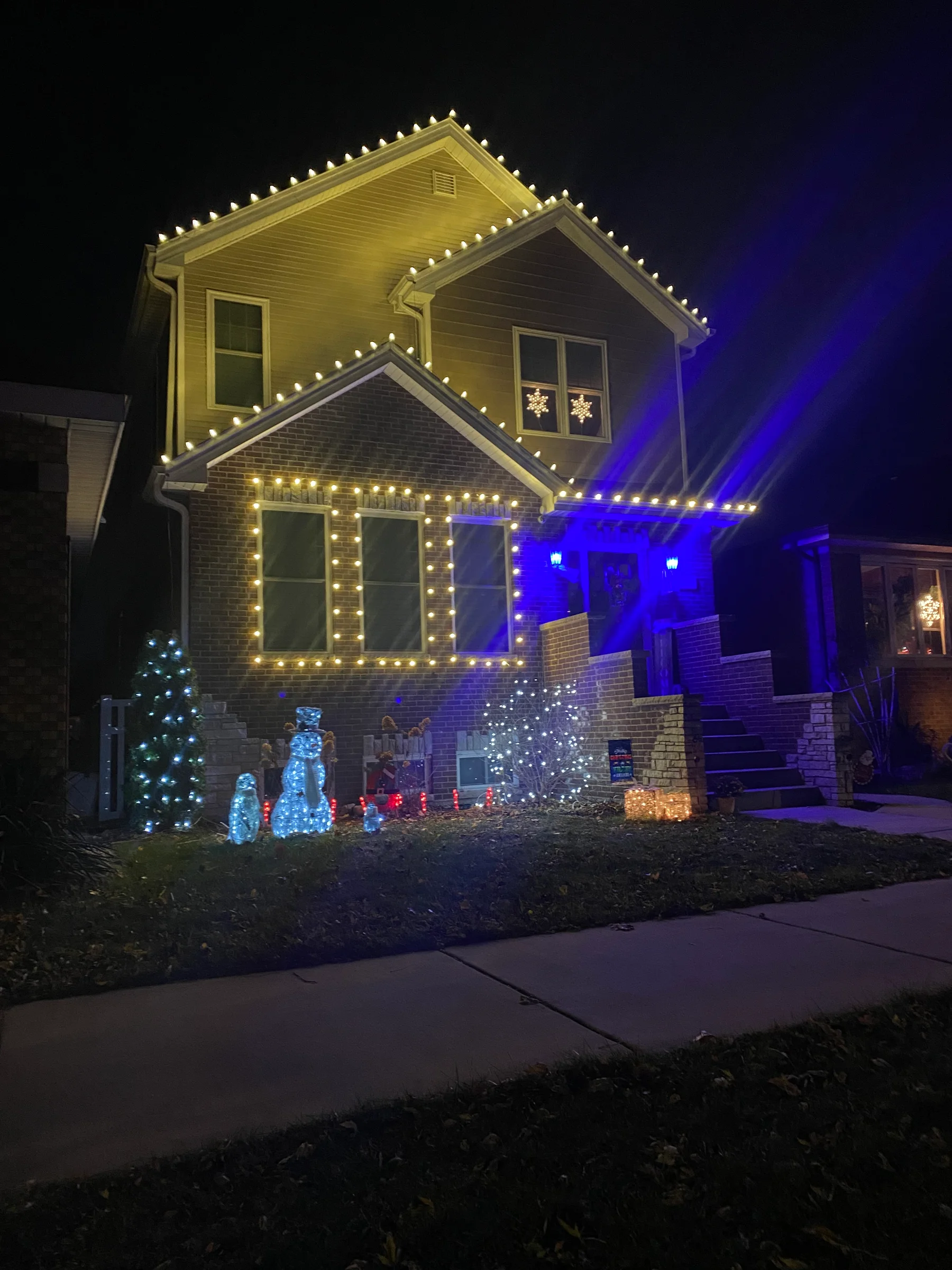 Two-story brick home with warm-white lights tracing multiple roof peaks, windows, entry, and front landscaping