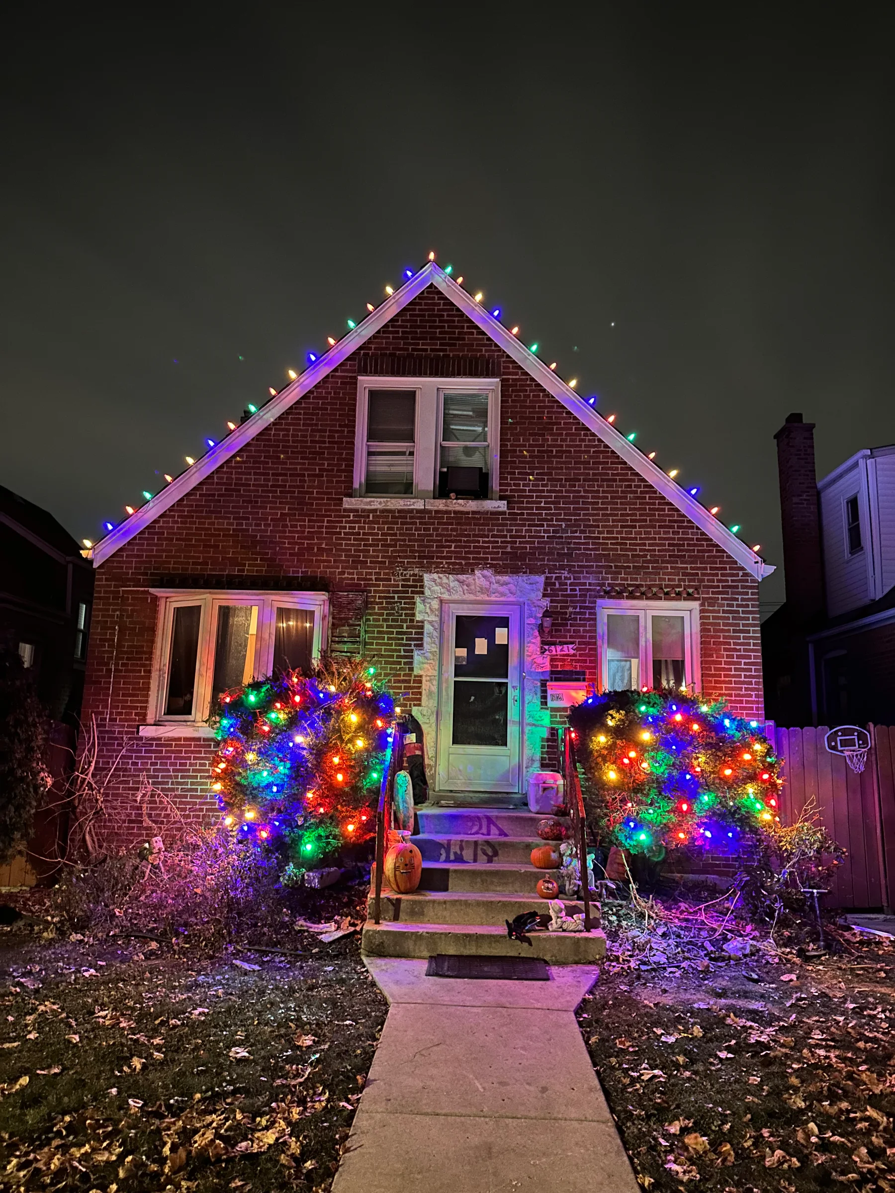 Compact brick home with colorful roofline lights and illuminated shrubs